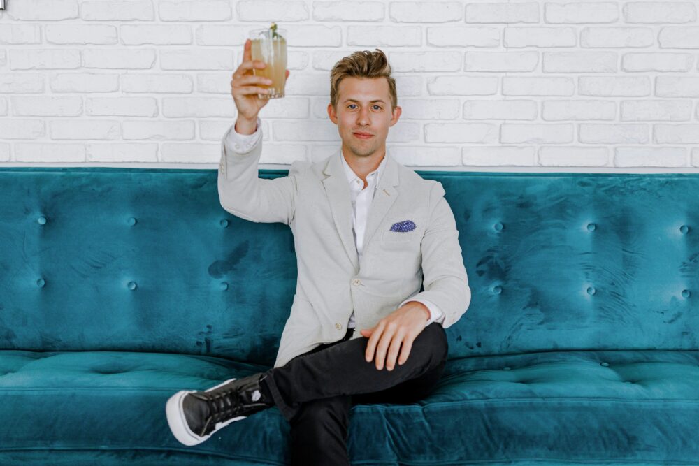 Young white rich man sitting on turquise sofa and cheering with a cocktail